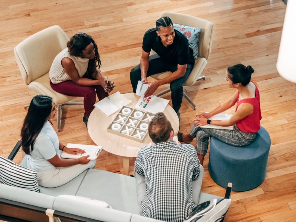 Group of five young professionals in discussion, gathered and seated around a round wooden coffee table.