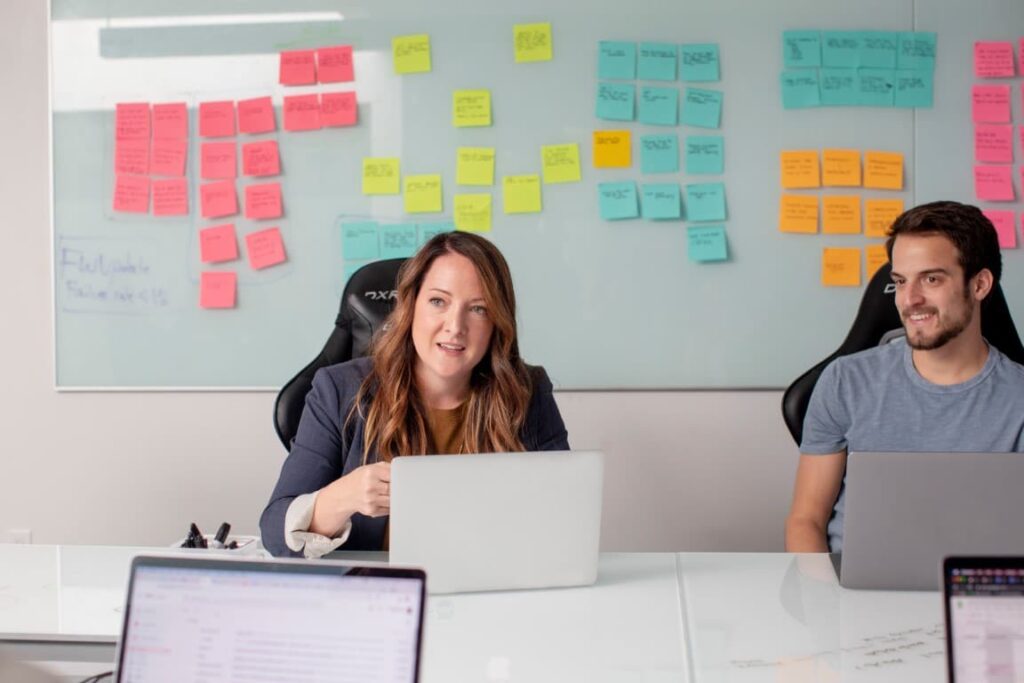 A professional woman and man sit at a white conference table with laptops in front of them in a group discussion.