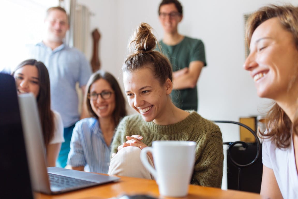 Group of six happy young professionals gathered around a laptop, watching the screen.