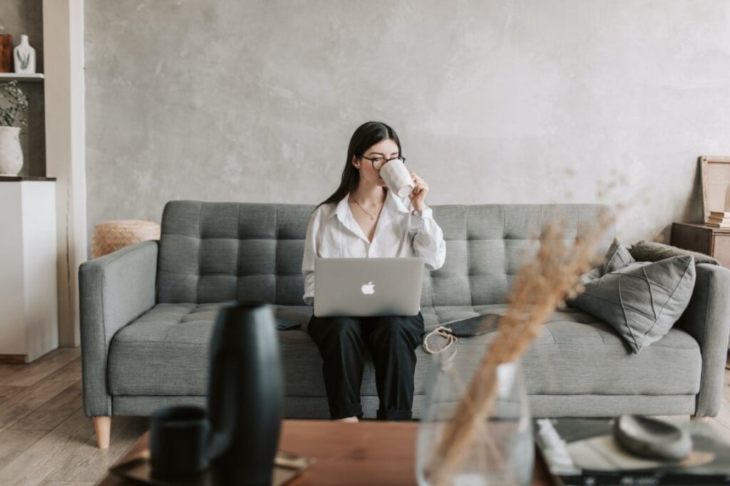 Woman with long dark brown hair wearing eyeglasses, a white long sleeve blouse and black pants drinks coffee and reads from her laptop on a gray couch.