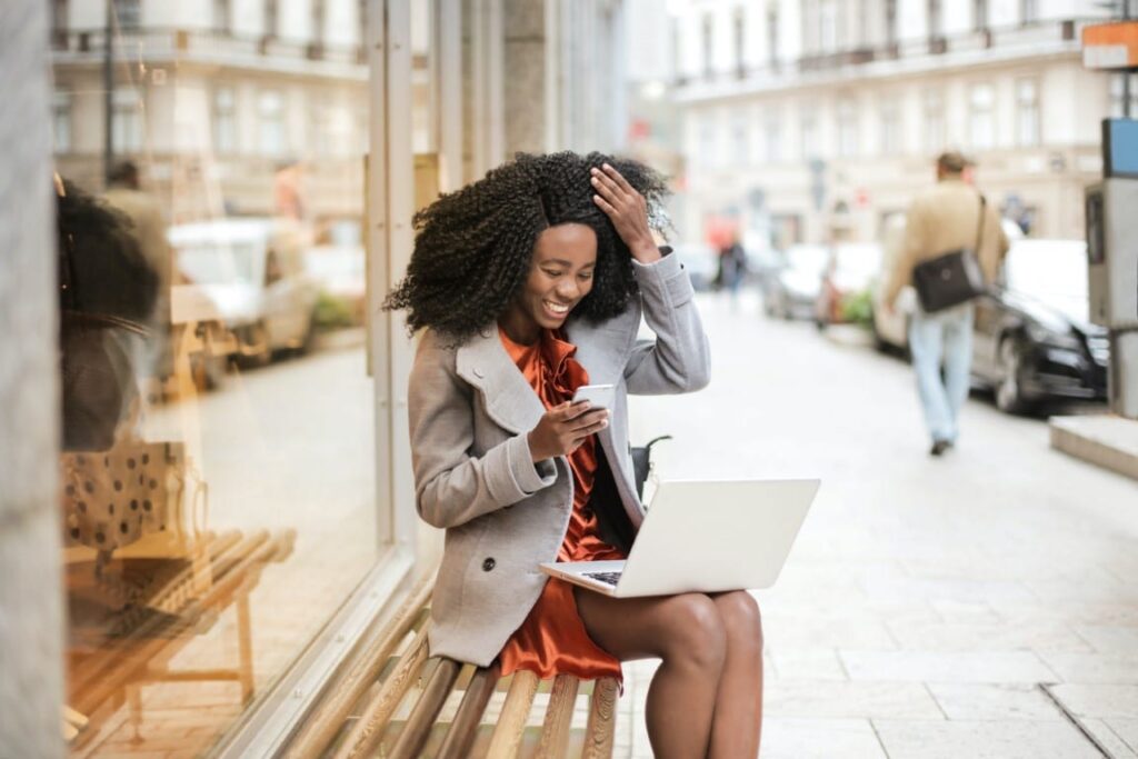 Happy, professionally dressed woman with dark curly hair sits on an outdoor city bench and looks at her phone in her right hand with a laptop situated in her lap.