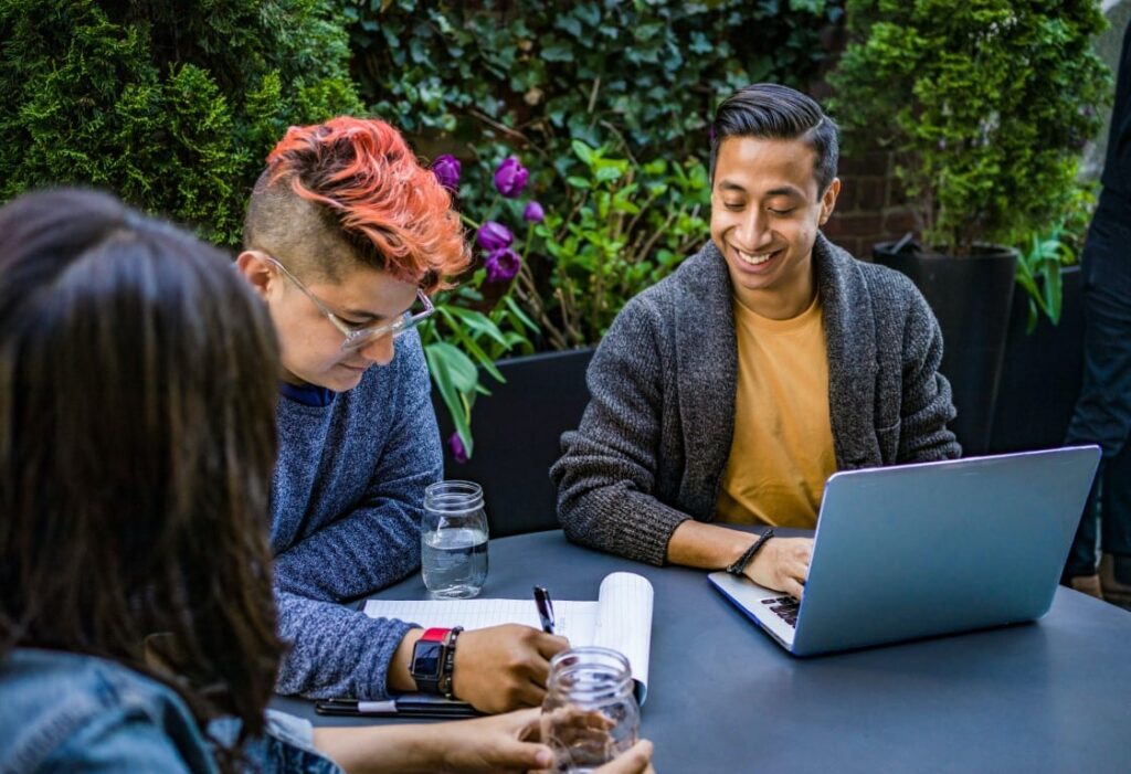 Group of three people sit at an outdoor table collaborating with an open laptop and notebook at hand.