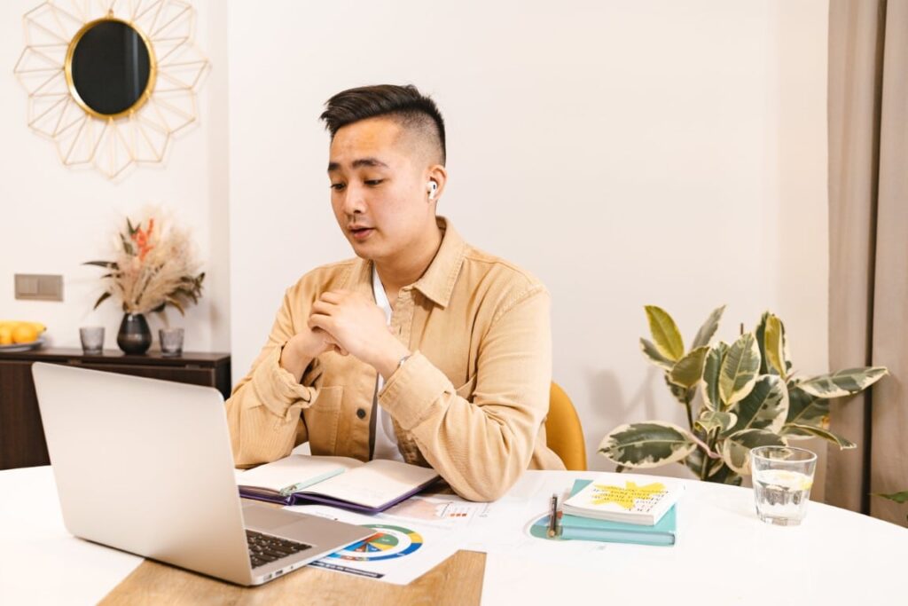 Man sitting at a table with an open laptop, papers and notebook is in the middle of a virtual meeting.