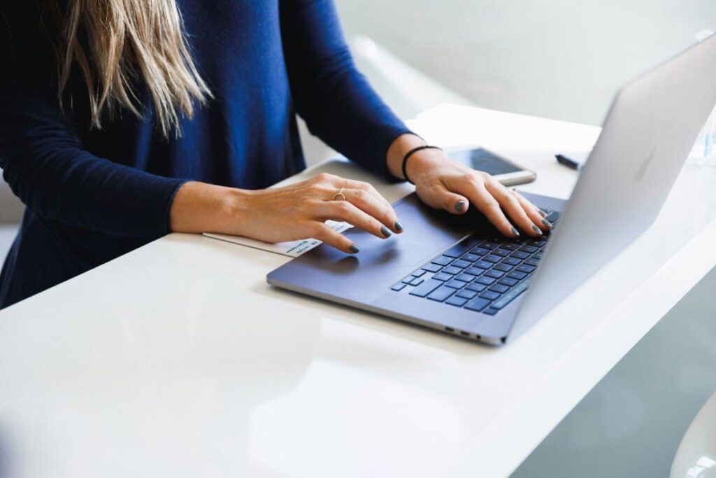 Woman with long hair, wearing a dark blue sweater sits at a white desk and uses her laptop.