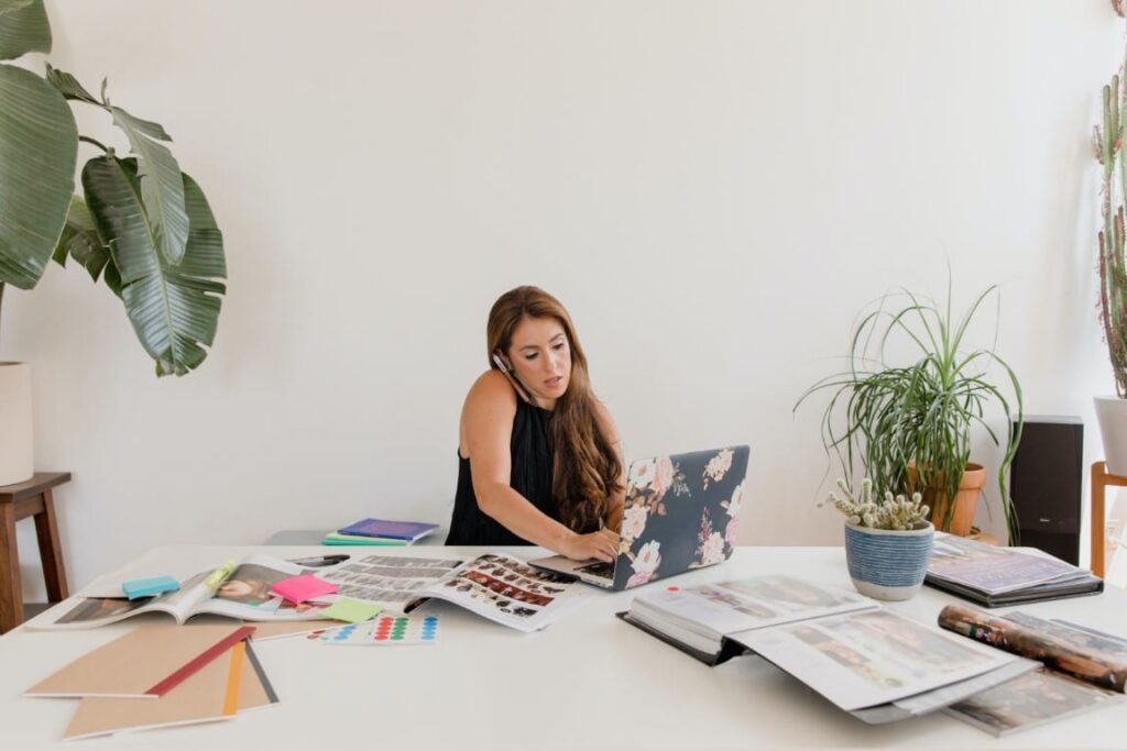 Business woman wearing a black sleeveless blouse talks on the phone and uses her laptop at a big desk covered with books, notebooks and magazines.