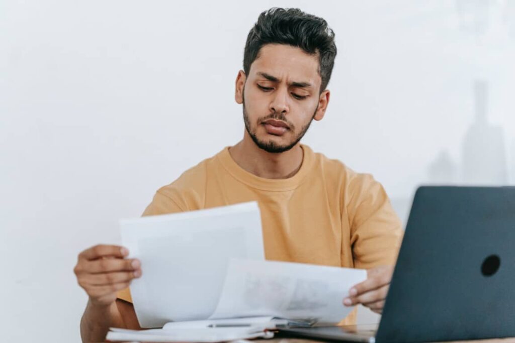 Man wearing a dark yellow t-shirt sits in front of his laptop with a concerned look on his face while reading papers in his hands.