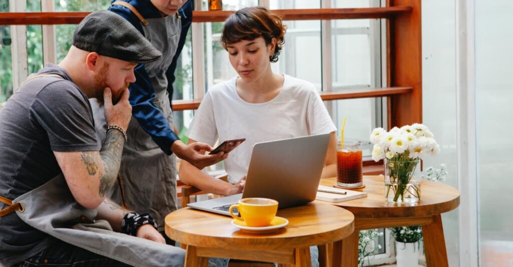 A man and a woman sit in front of two small coffee tables while looking at the screen of a phone that’s held by a man that stands between them.