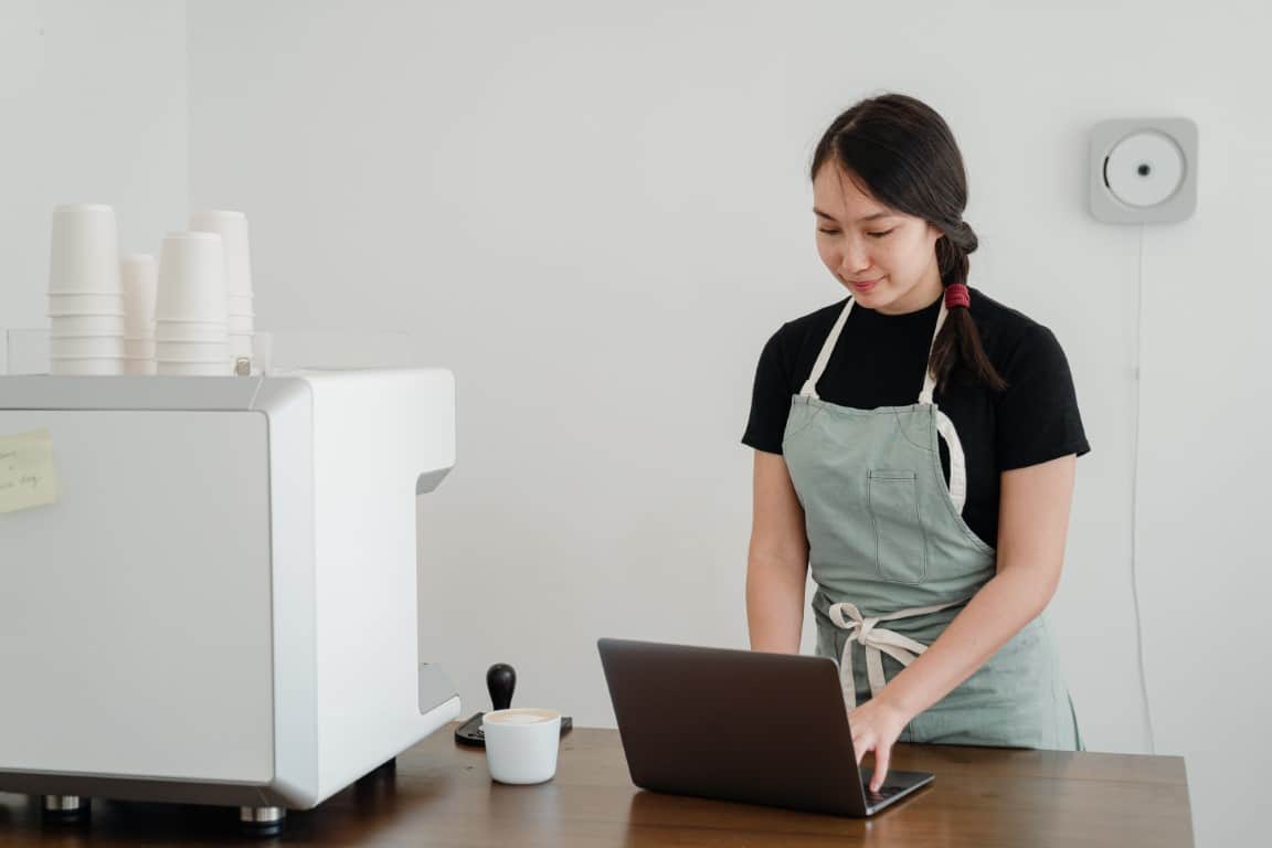 Female coffee shop business owner stands behind the counter typing on her laptop.