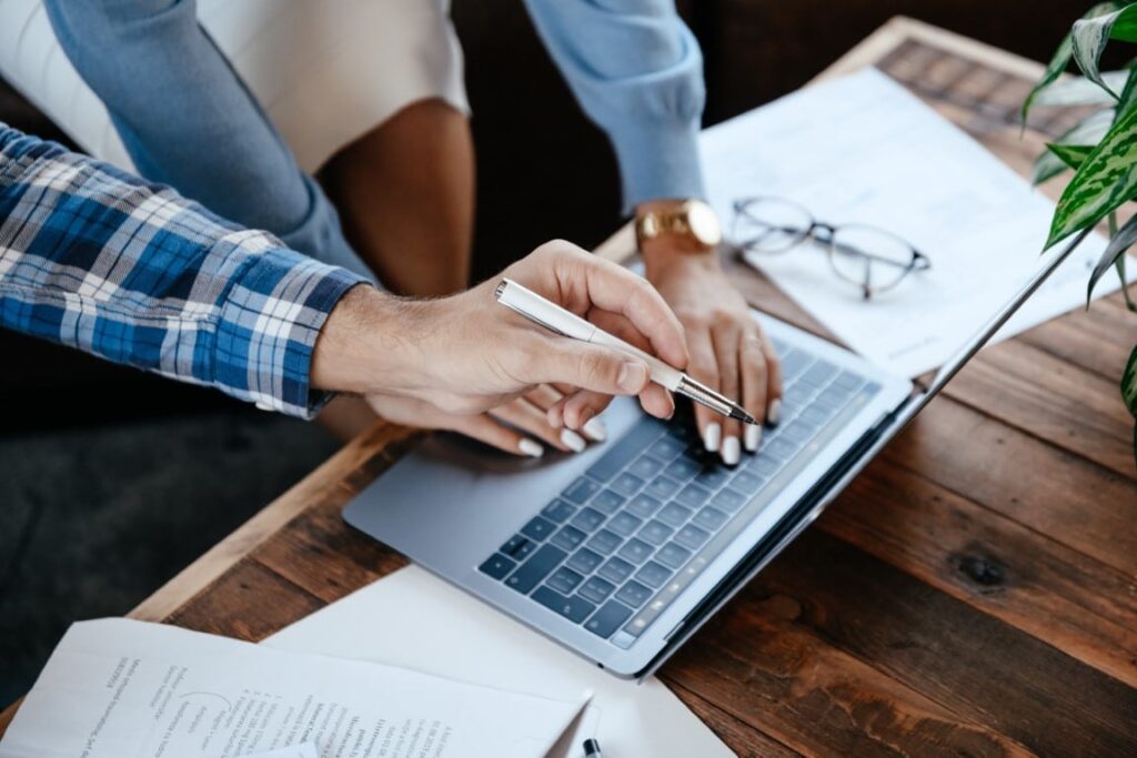 A man wearing a blue and white plaid long sleeve shirt uses a pen in his hand to point at a laptop screen while a woman wearing a blue long sleeve blouse sits at the laptop with her hands on the keyboard.