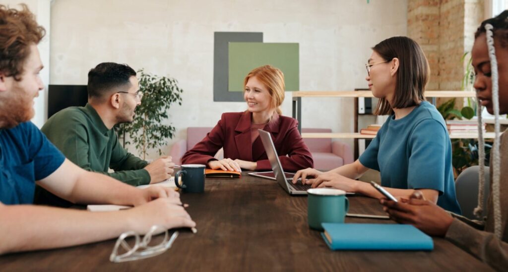 A group of five sit at a long wooden table in discussion with laptops, notebooks and coffee.