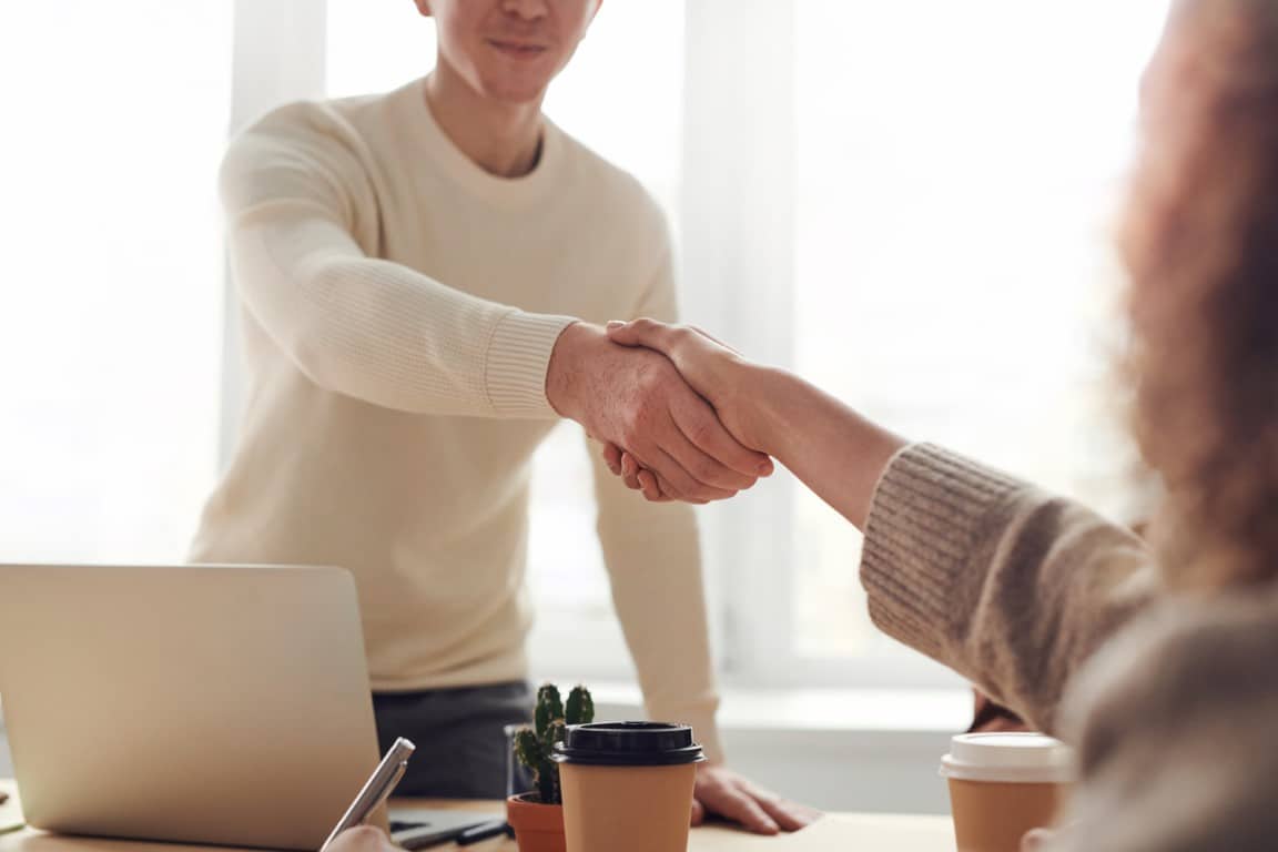 Man behind a desk wearing a white sweater stands up to shake the hand of a woman sitting on the other side of the desk.