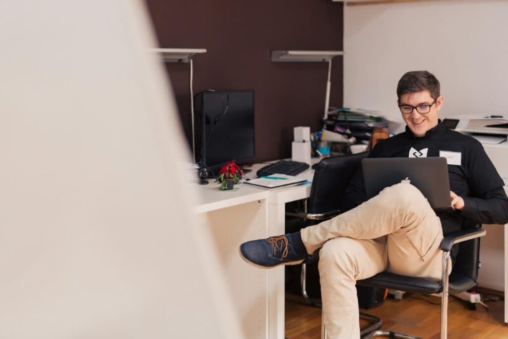 Man wearing a black shirt, khaki pants and dark frame eyeglasses uses his laptop situated on his lap.