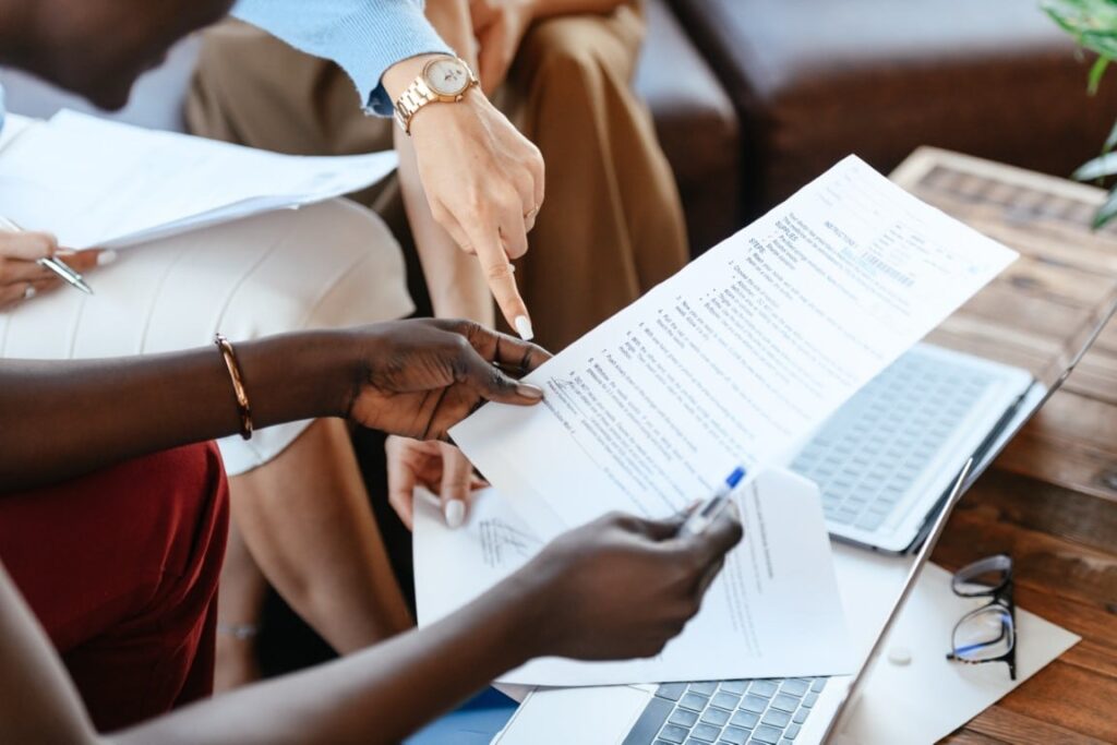 Three business women sit in front of a wooden coffee table with paperwork and two open laptops in front of them.