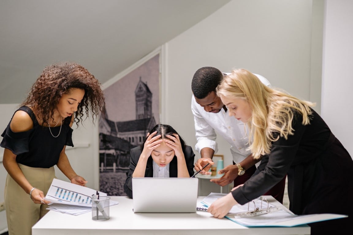 Group of four professionals look concerned while searching for information in paper files and on a laptop.