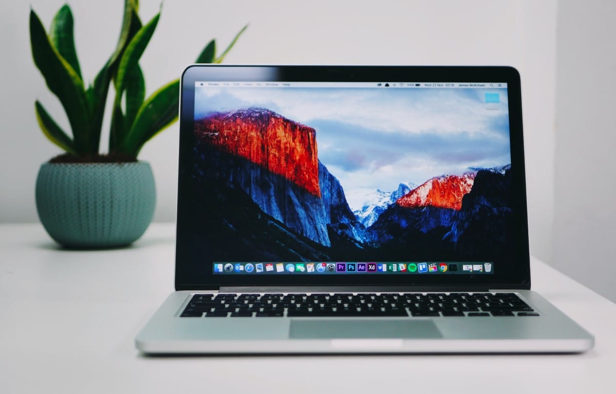 An open, powered-on laptop, with a leafy green plant planted in a short turquoise planter behind it, sits on a white table.