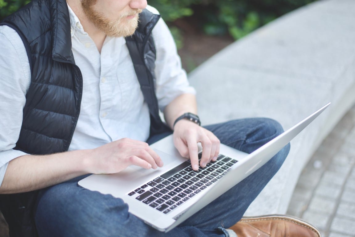 Man with short beard wearing a light gray button down shirt, black puffy vest and jeans sits on a concrete bench and types on his laptop, which is held in his lap.