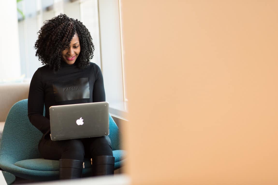 Woman with curly black hair wearing a black long sleeve shirt, black pants and black boots sits on a teal chair typing on her laptop, which is held in her lap.
