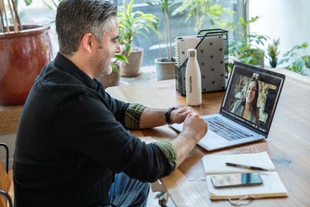 A man wearing a black long sleeve collared shirt sits at a wooden table in a virtual meeting with a woman displayed on his laptop screen.