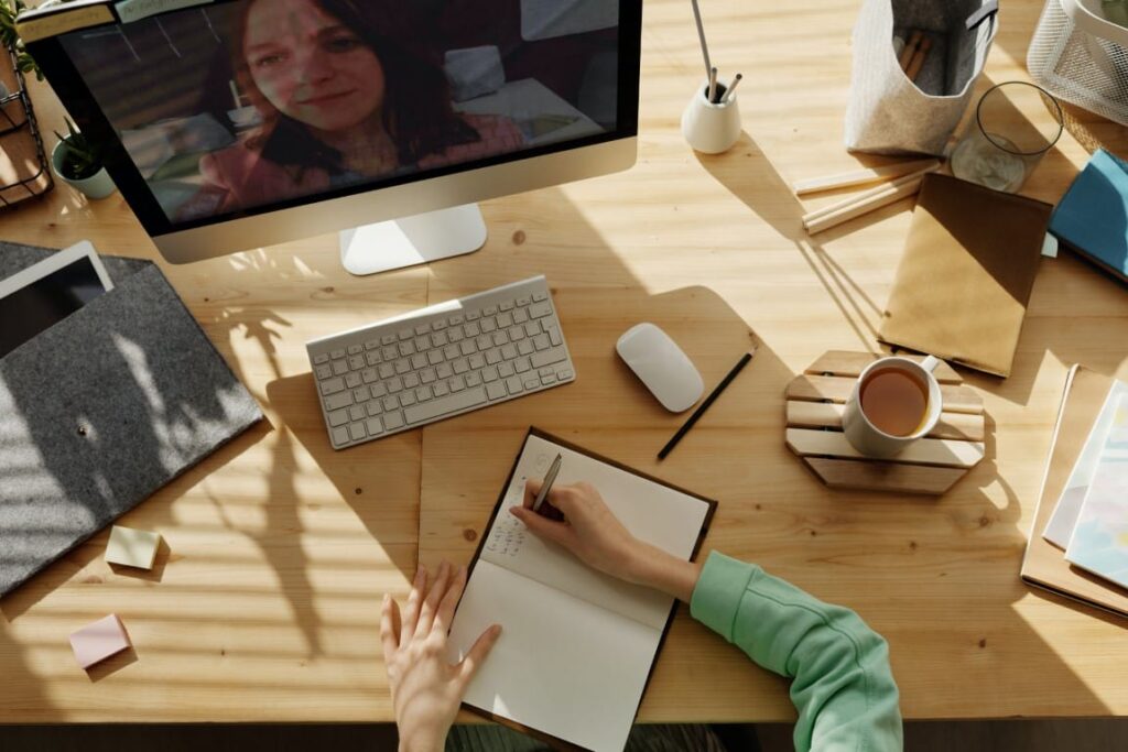 A top view of a wooden desk with two women in a virtual meeting. One woman writes notes in a notebook at the wooden desk and the other woman is displayed on the large computer monitor.