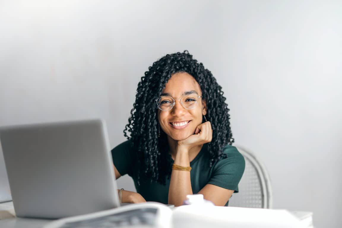 A young smiling woman wearing a dark green short sleeve shirt sits at a table with books and a laptop in front of her.