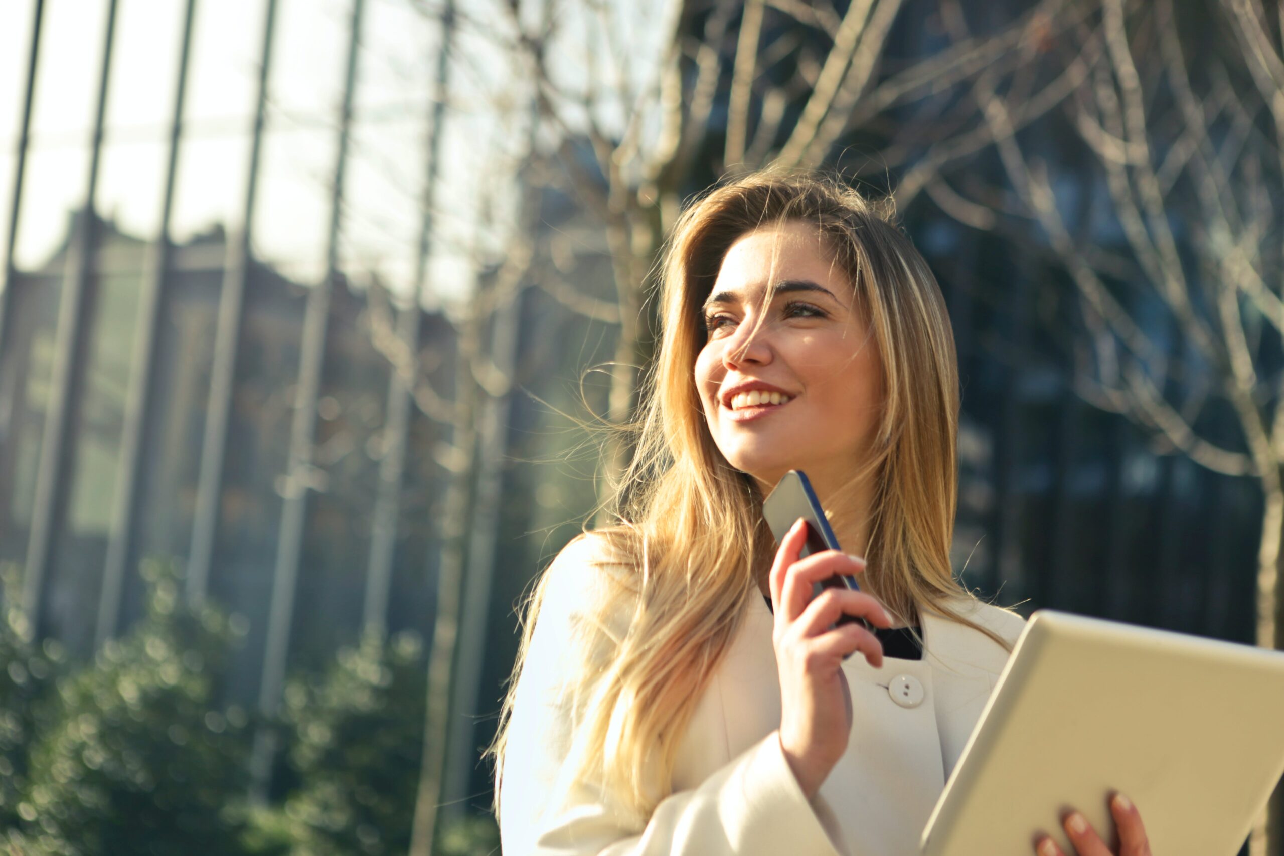 A woman with long blonde hair wearing a white peacoat looks in the distance with a smile on her face, holding a cell phone in her right hand and a tablet in her left hand.