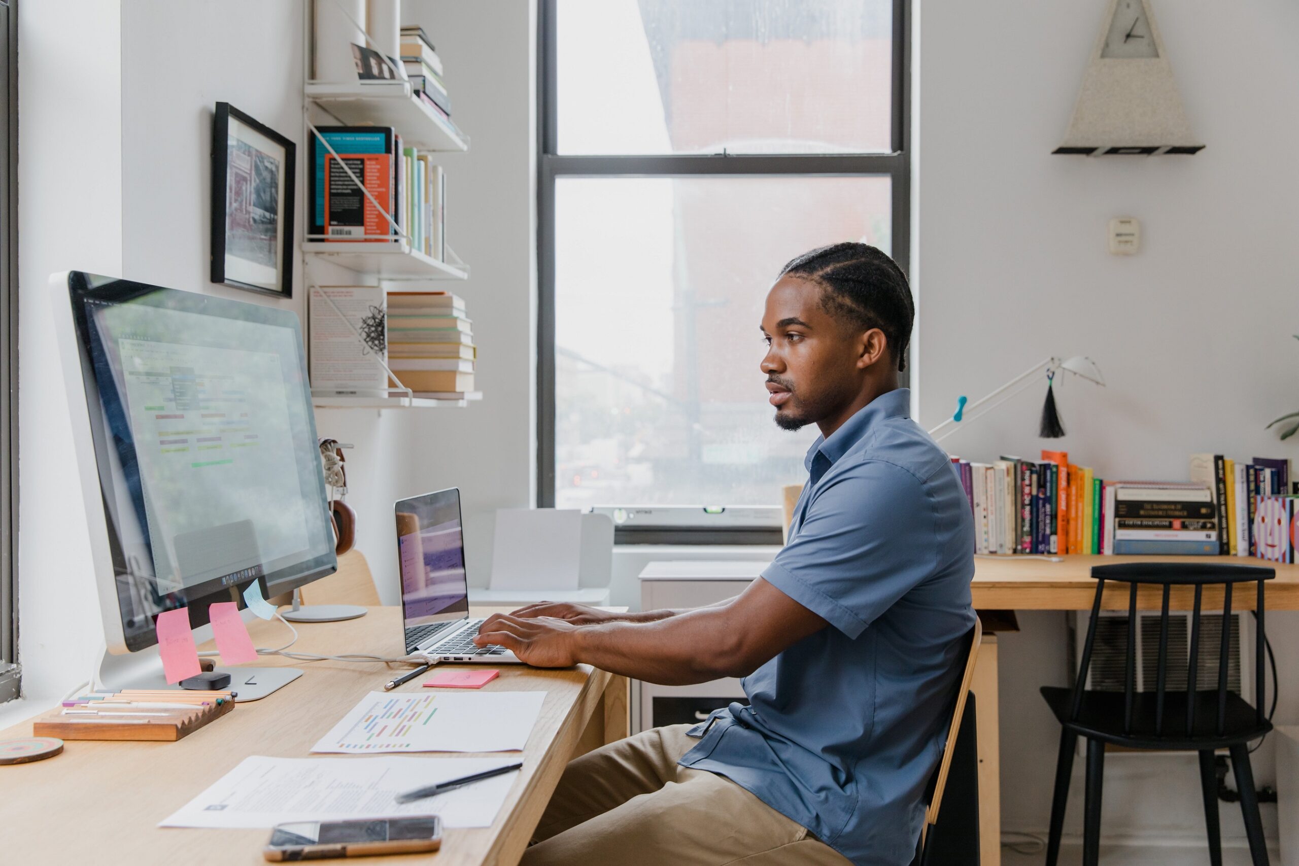 A man wearing a blue collared t-shirt sits at his desk typing on his laptop while looking at a larger computer monitor.