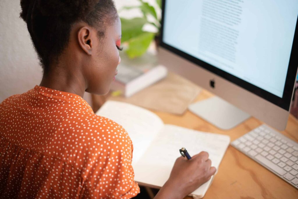 A woman wearing an orange blouse with white polka dots writes in a notebook at her desk with a desktop computer and keyboard.