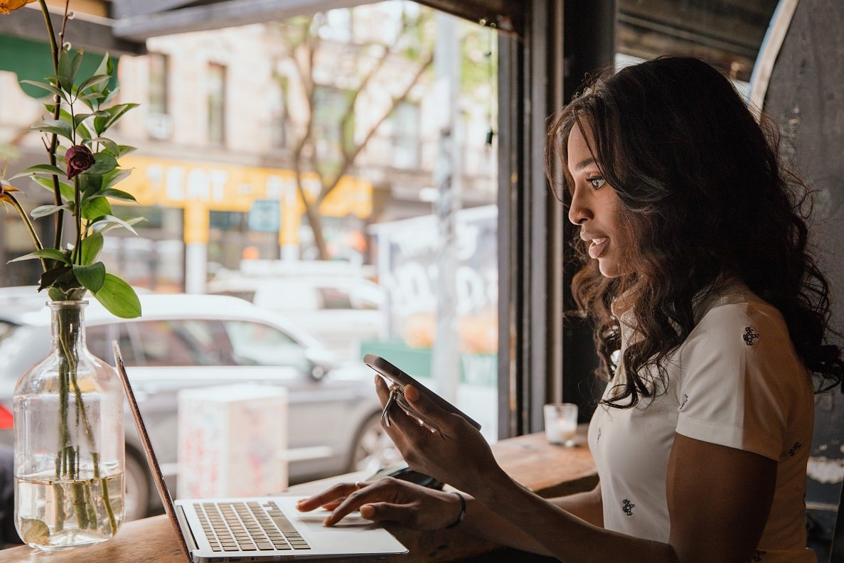 A woman sitting at a window counter uses her laptop while holding her cell phone in her left hand.