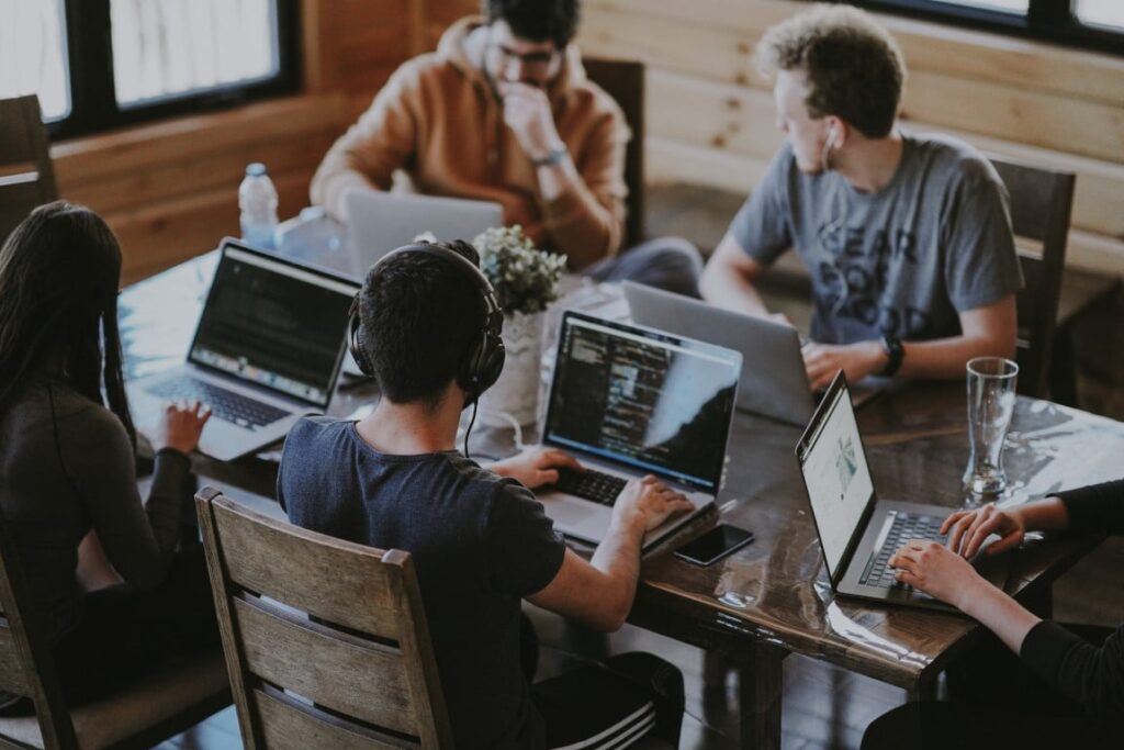 A group of five young adults sit together at a table with open laptops in front of each person.
