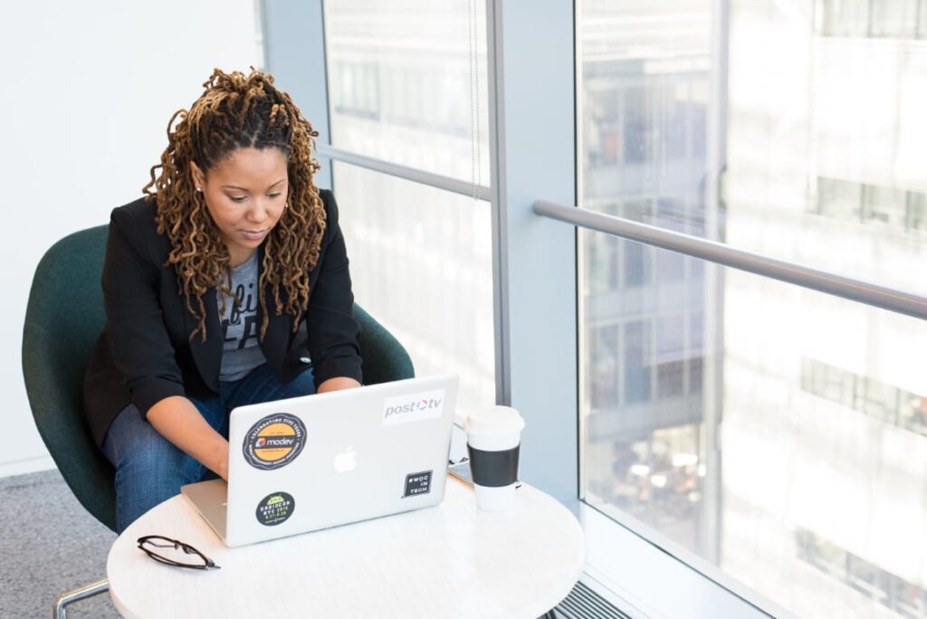 a woman wearing a black blazer and jeans sits near the window of a high-rise building typing on her laptop.