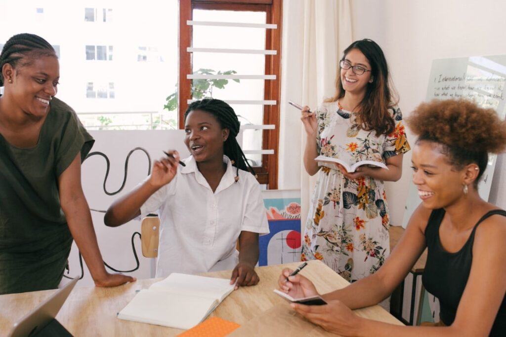 A group of 4 happy businesswomen surround a work table in discussion and writing in notebooks.
