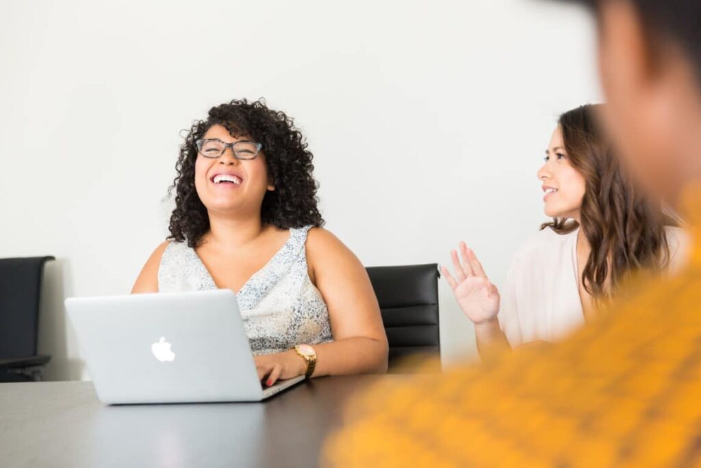 Three business women sit at a table together in conversation. One woman with dark curly hair and black rimmed glasses sits in front of a laptop.