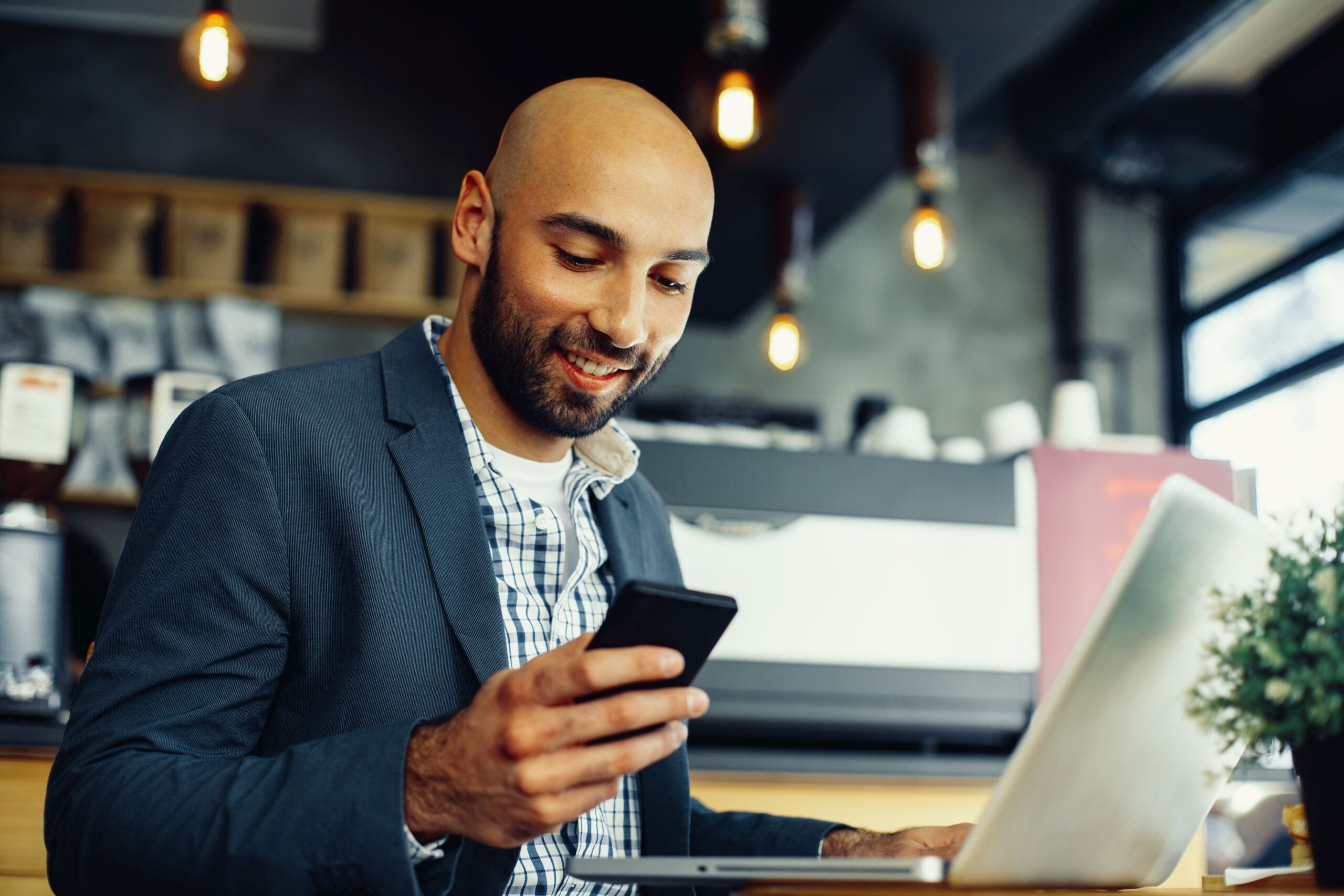 AI Chatbot and agents, image of a male professional in a coffee shop looking at his phone with his laptop opened in front of him.