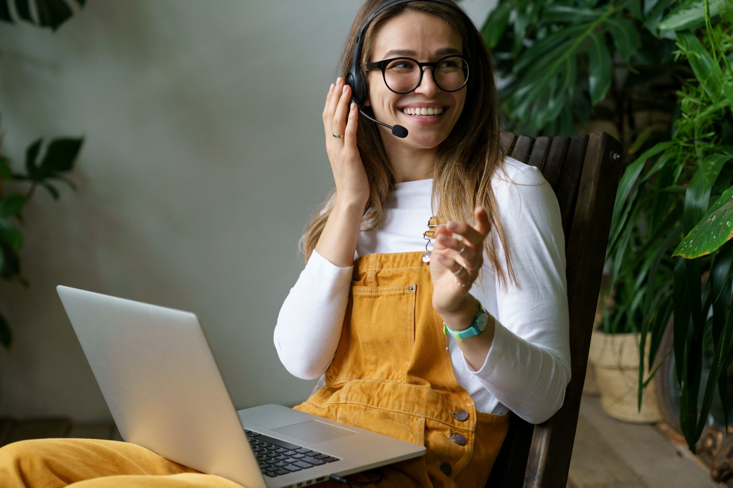 AI Chatbot and agents, image of a young woman wearing a white long sleeve shirt, dark yellow overalls, and a headset smiles with a laptop on her lap.