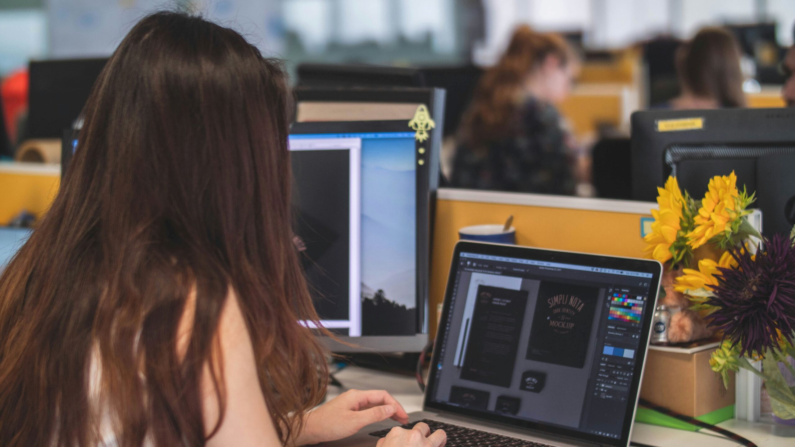 Facebook ad graphics, image of a woman designing graphics at a desk with a laptop, large computer monitor and a vase of sunflowers