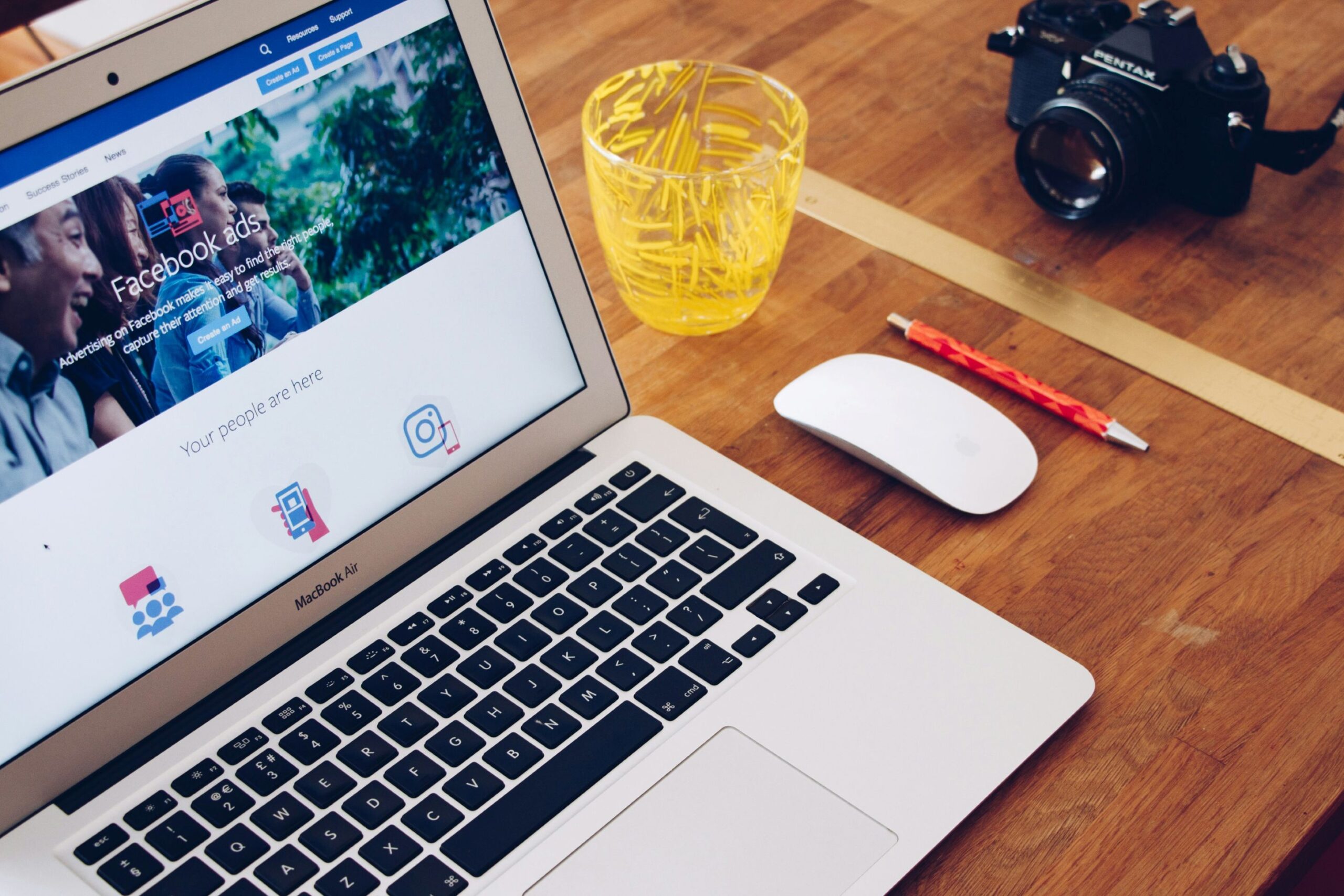 Facebook ad graphics, image of an open laptop with Facebook Ads webpage on the screen with a yellow cup, white computer mouse, red pen and camera next to the laptop.