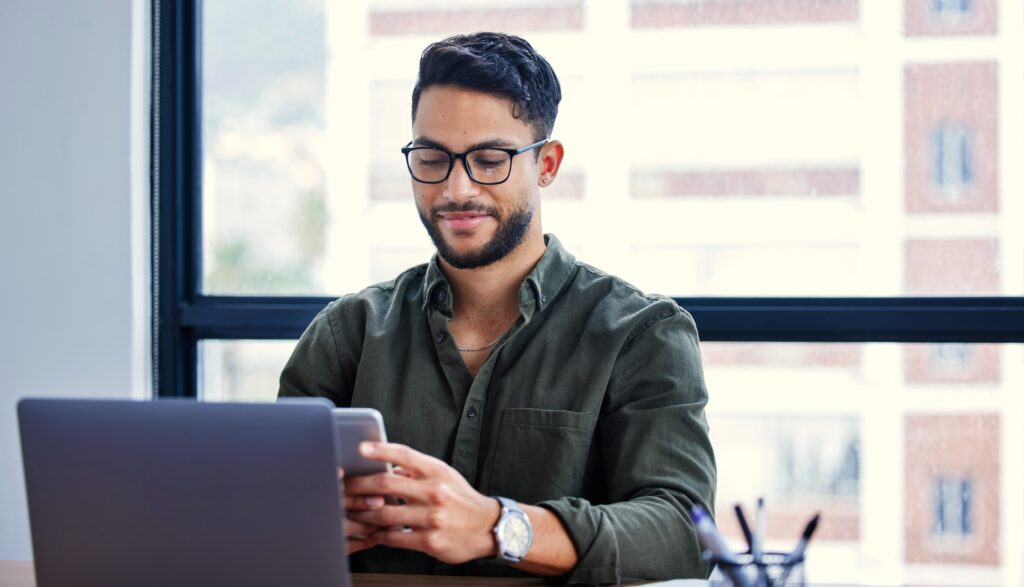 Important Meta Update, image of a man wearing dark rim glasses and a dark green button down shirt sitting at a desk with his smartphone in his hands and his laptop in front of him.