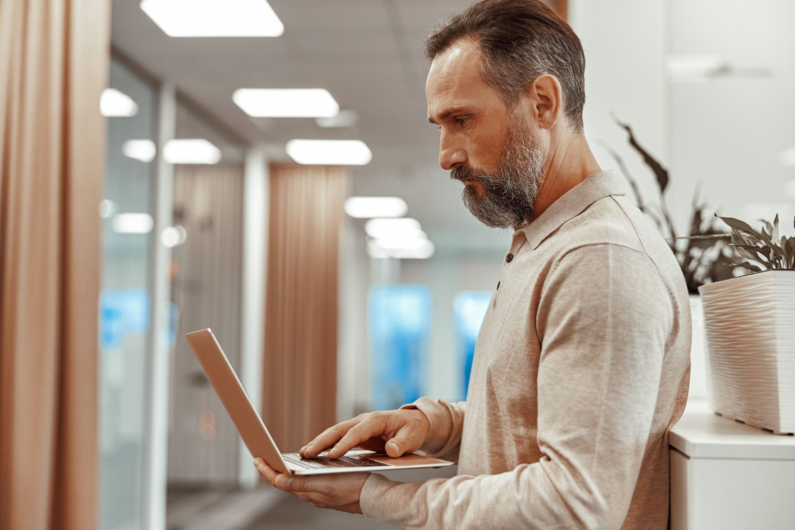 The Best EHR Systems for Small Practices, image of a man wearing a beige button-down collared shirt stands and uses his laptop in his left hand with his right hand on the keyboard.