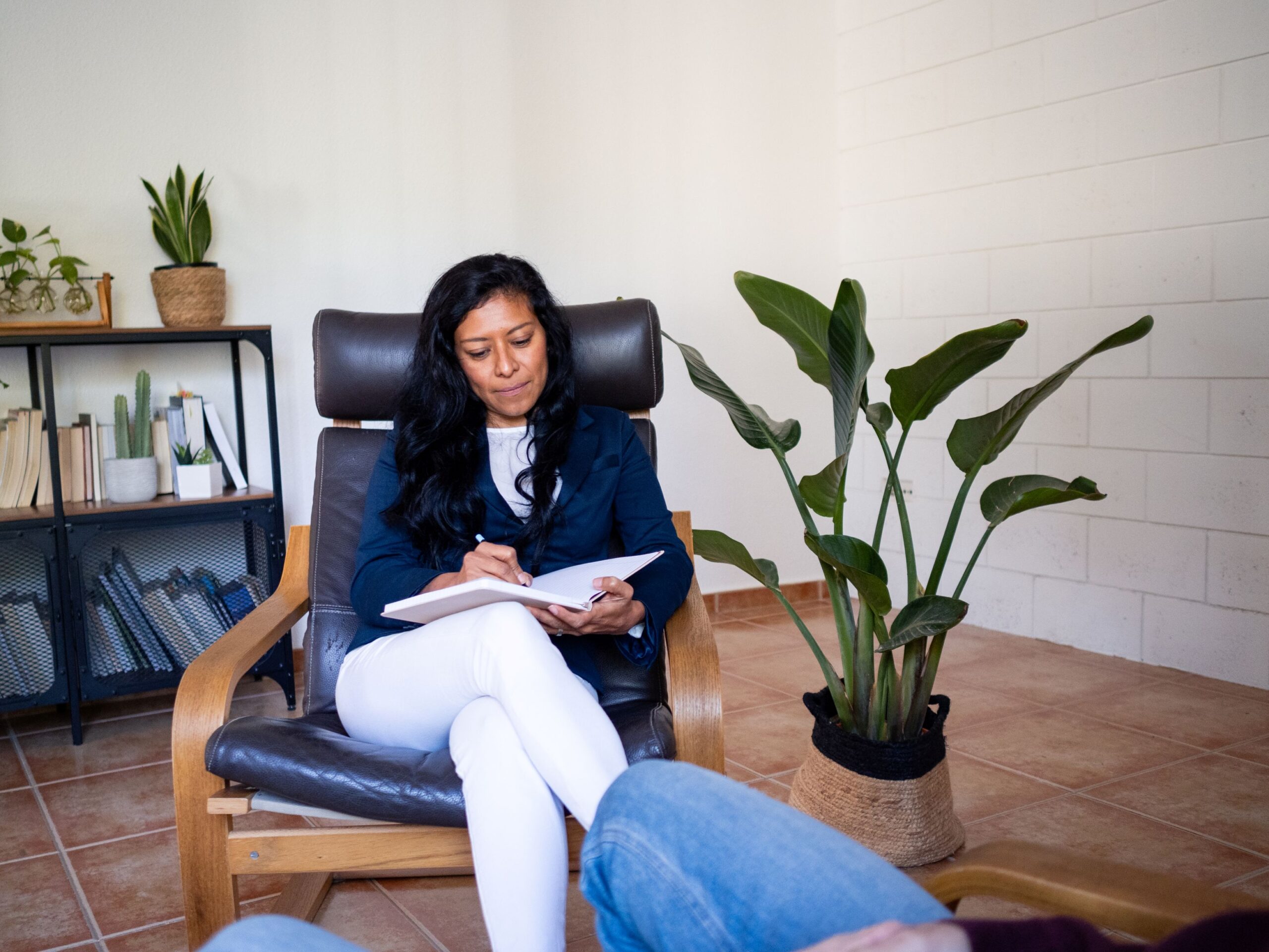 The Best EHR Systems for Small Practices, image of a woman therapist with long dark hair, a blue jacket and white pants sitting in a leather arm chair writes in her notebook with a patient sitting across from her.