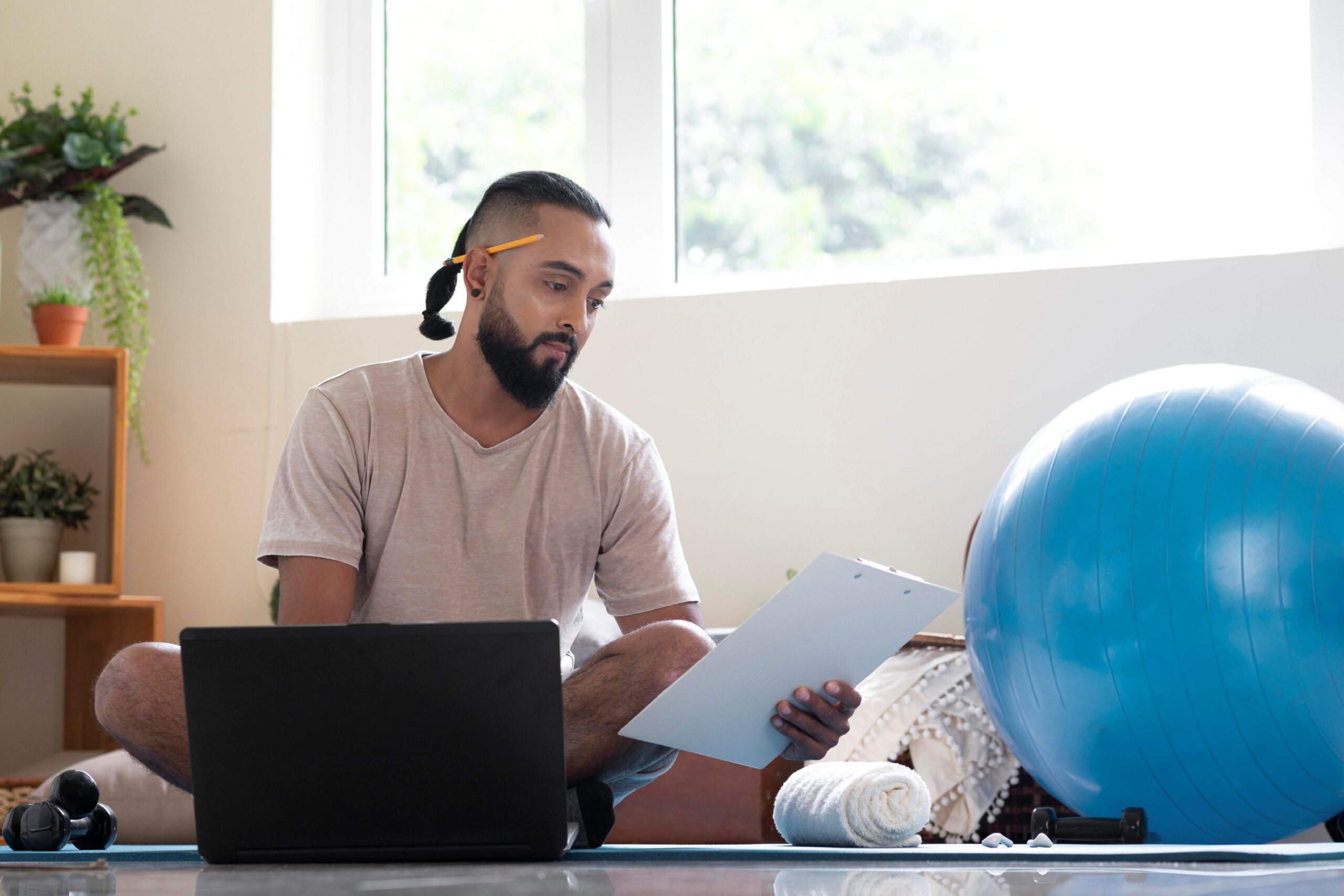 Three Things Your Clinic Can Do Right Now to Boost SEO, image of a physical trainer sitting on the floor in front of his opened laptop with a clipboard in his left hand and exercise equipment around him.