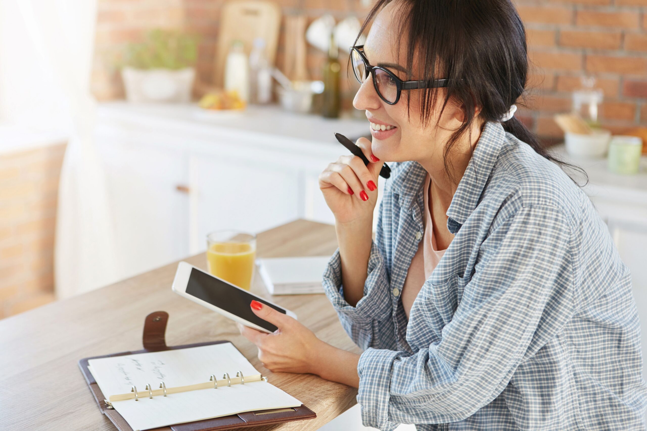 Three Things Your Clinic Can Do Right Now to Boost SEO, image of a woman sitting at a kitchen counter with her smartphone in her left hand and a pen in her right hand with a planner book and glass of orange juice on the counter.