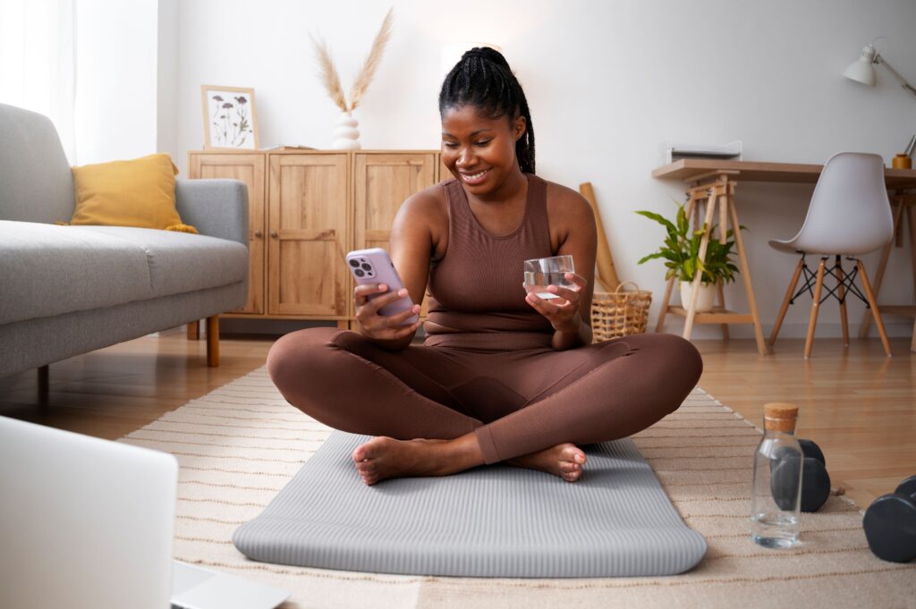 What Does It Mean to Be Everywhere Potential Patients Are Searching, image of a woman in a brown yoga outfit sitting on a yoga mat looking at her smartphone in her right hand with a glass of water in her left hand.