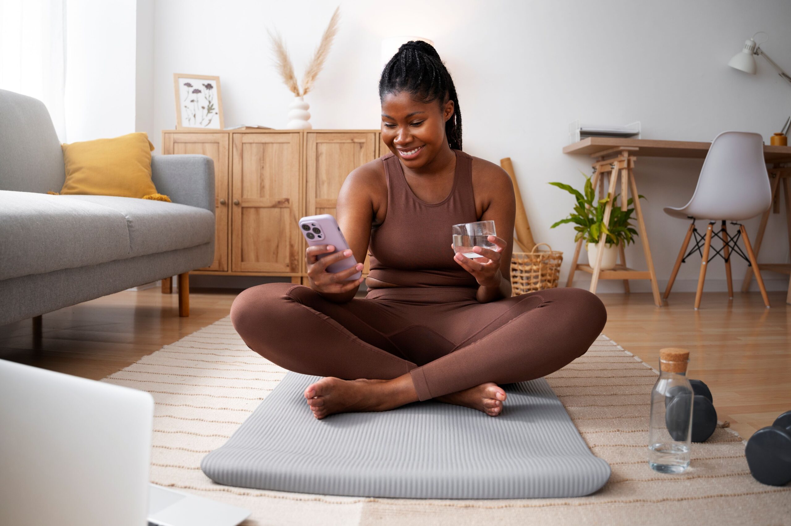 What Does It Mean to Be Everywhere Potential Patients Are Searching, image of a woman in a brown yoga outfit sitting on a yoga mat looking at her smartphone in her right hand with a glass of water in her left hand.