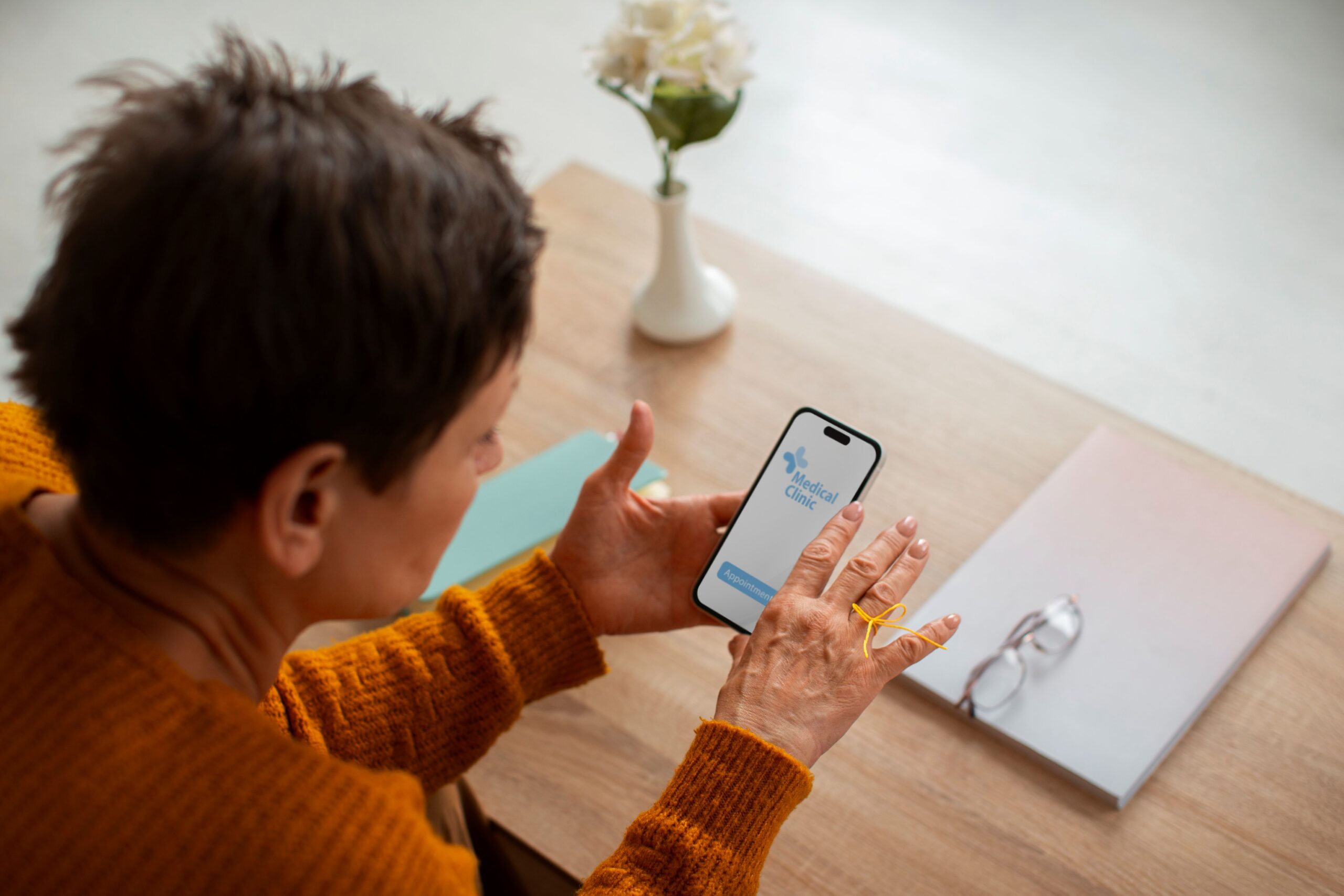 What Does It Mean to Be Everywhere Potential Patients Are Searching, image of a woman wearing an orange sweater sitting at a wooden table with a flower vase and notepad uses her smartphone with a medical clinic application opened.