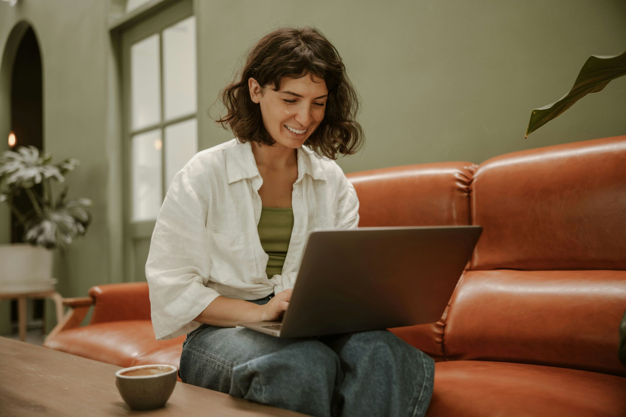 Wix Groups, image of a smiling woman wearing a white knotted blouse with a green undershirt and jeans sits on an orange leather couch with a laptop in her lap.