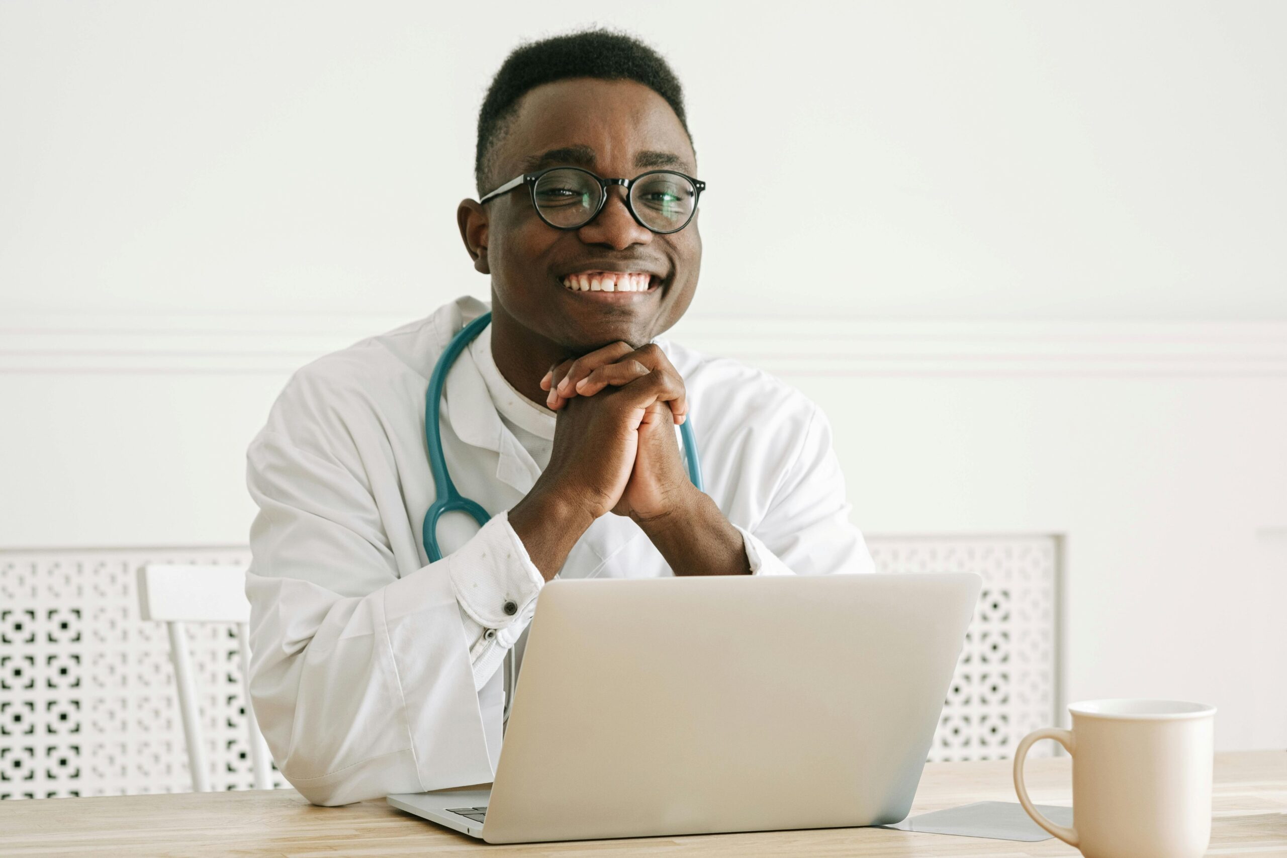 Wix vs WordPress, image of a smiling doctor sitting at a table with a white coffee cup and open laptop in front of him with his hands held together under his chin.
