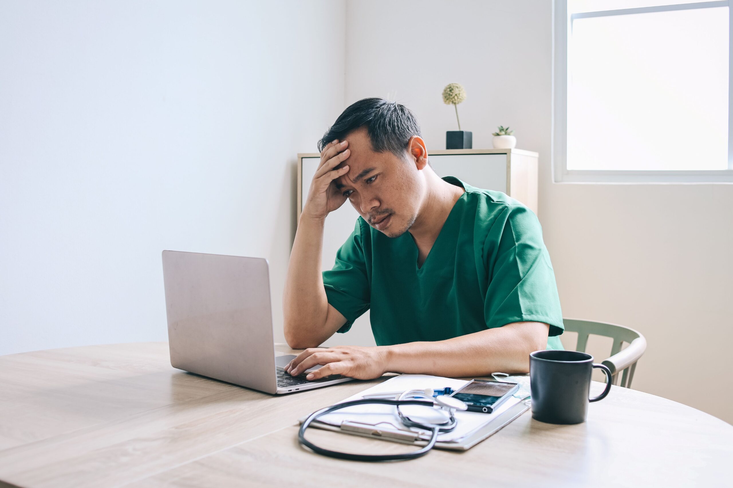 WordPress hacked, image of a frustrated man in green medical scrubs sitting at a wooden table in front of his laptop, clipboard, coffee cup and stethoscope with his right hand over his forehead.