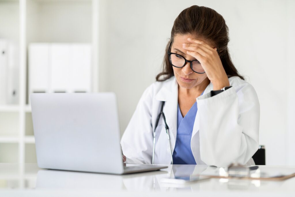 WordPress hacked, image of a frustrated doctor with long brown hair and black frame glasses sits in front of her laptop at a table with her left hand over her forehead.