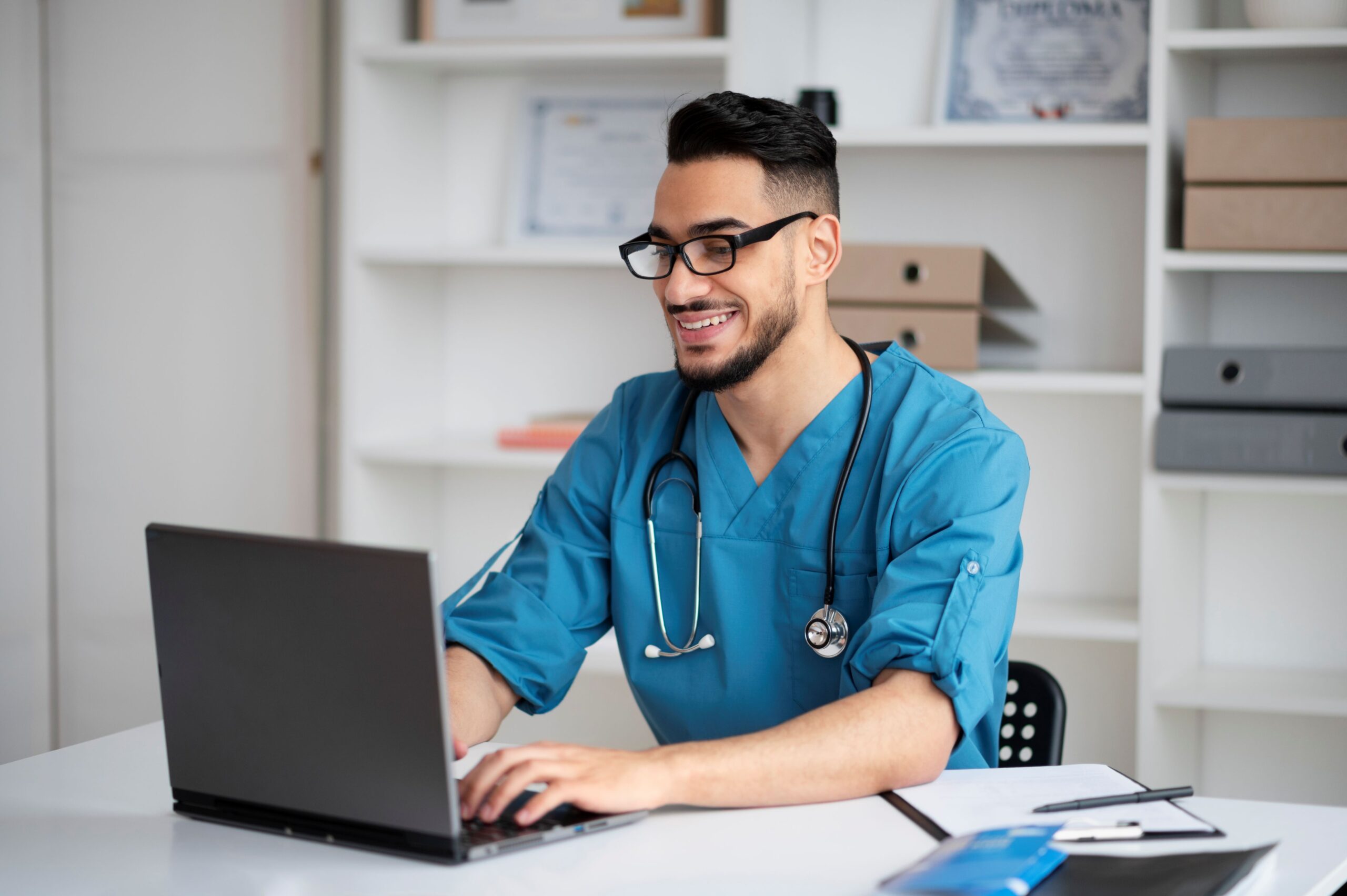 Youtube policies, image of a smiling male doctor in blue scrubs and a stethoscope around his neck sits at a white desk and types on a laptop.