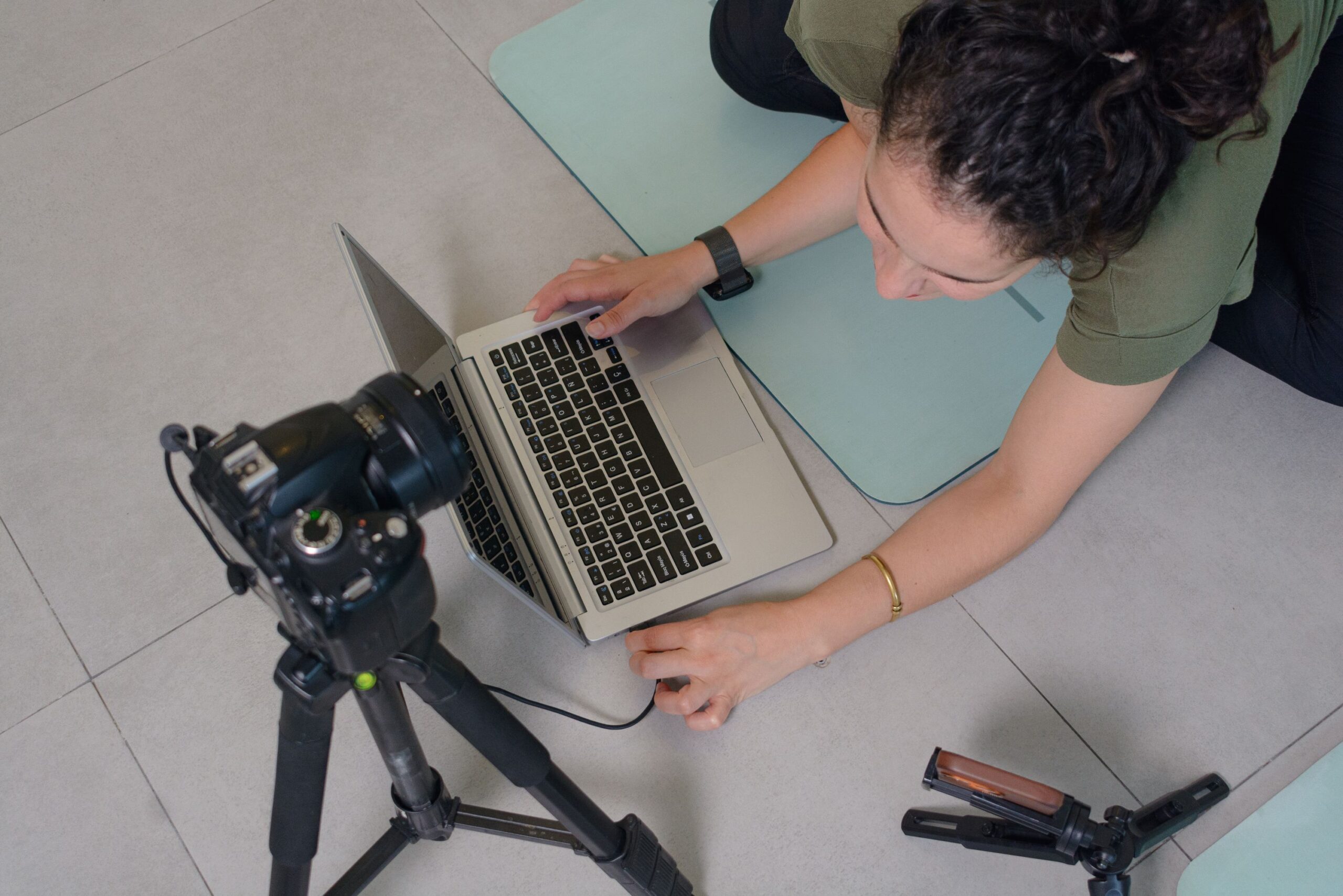 Youtube policies, image of a woman on a blue yoga mat plugs her camera, mounted on a camera stand, into her laptop that’s placed on the floor in front of her.